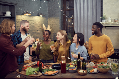 Multi-ethnic group of smiling young people enjoying dinner together standing at table in modern interior and holding wine glasses, copy space